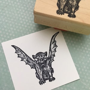 May include: A black and white rubber stamp with a gargoyle design. The gargoyle has wings and a menacing expression.