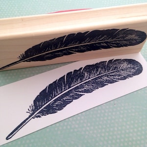 May include: A wooden stamp with a black and white image of a feather. The stamp is mounted on a wooden block. The feather is a simple design with a few lines to represent the quill.