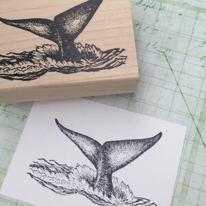 May include: Black and white illustration of a whale tail splashing in the water. The image is printed on a wooden block.