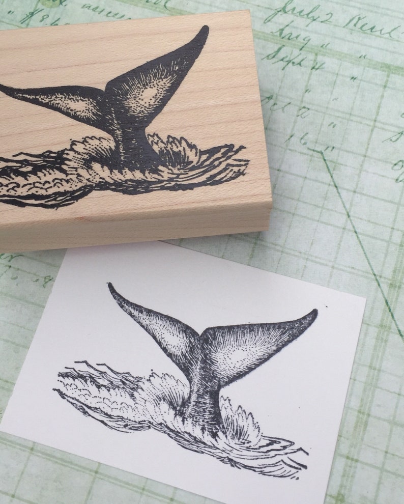 May include: Black and white illustration of a whale tail splashing in the water. The image is printed on a wooden block.