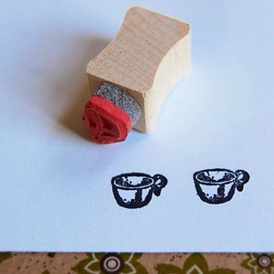 Könnte beinhalten: Ein Stempel aus Holz mit einem roten Gummidesign von zwei Teetassen. Der Stempel befindet sich auf einer weißen Oberfläche.