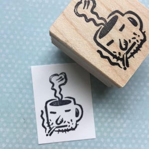 May include: A black and white rubber stamp featuring a cartoon illustration of a coffee mug with a face and steam rising from the top. The mug is holding a cigarette in its mouth.