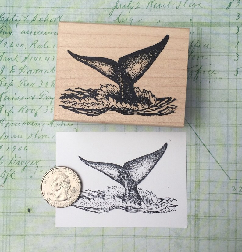 May include: A rubber stamp with a black and white illustration of a whale tail splashing in the water. The stamp is made of wood and has a rectangular shape.