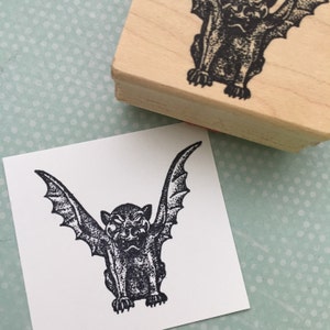 May include: A black and white rubber stamp featuring a gargoyle with wings. The gargoyle is depicted with a fierce expression and is in a standing position with its wings spread out.