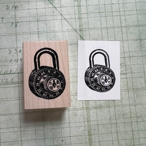 May include: A wooden rubber stamp with a black and white image of a combination padlock. The padlock has a dial with numbers from 0 to 40.