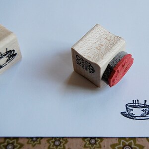 Peut inclure: Deux tampons en bois avec une image en noir et blanc d'une tasse &agrave; th&eacute;. Le tampon de gauche porte le texte "100 Proof Press" sur le c&ocirc;t&eacute;. Le tampon de droite a un tampon encreur rouge.