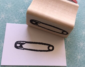 May include: A wooden stamp with a black and white image of a safety pin. The stamp is on a white card with a black and white image of a safety pin.
