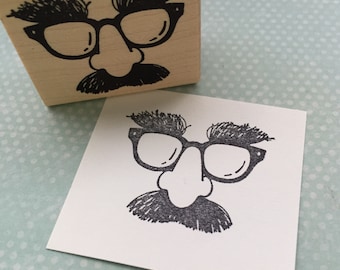 May include: A black and white rubber stamp with a cartoon face wearing glasses, a large nose, and a mustache. The stamp is mounted on a wooden block.