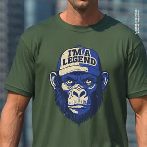 May include: Olive green t-shirt featuring a blue and beige illustration of a chimpanzee wearing a blue baseball cap. The cap has the text "I'M A LEGEND" in white letters.