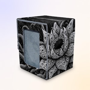 May include: A black and white cube-shaped storage box featuring a detailed illustration of a dark, multi-petaled flower covered in numerous eyes and sharp teeth, with a plain gray rectangular panel on the front side of the container.