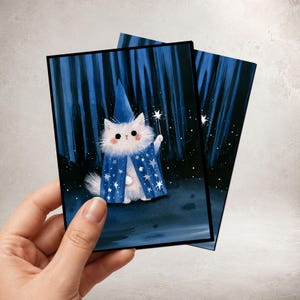 May include: A rectangular greeting card featuring an illustration of a fluffy white cat wearing a blue wizard hat and a matching blue cape decorated with white stars. The cat holds a small white star-tipped wand against a dark blue forest background.