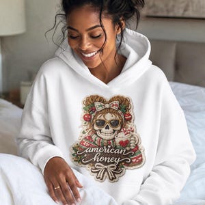 May include: A white hooded sweatshirt featuring a central graphic of a skull wearing sunglasses, a bow, and a scarf. The skull holds a mug with a candy cane. The text "american honey" is written in cursive script below the skull, with a bow at the base.