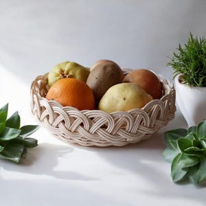 Może przedstawiać: Pleciony beżowy kosz wypełniony świeżymi owocami, w tym pomarańczą, gruszkami i kiwi. Kosz ma dekoracyjny pleciony wzór. Zielone rośliny i biała doniczka w tle.