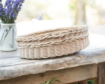 Puede incluir: Una cesta de mimbre tejida de color marrón claro con un borde trenzado. La cesta de forma ovalada se encuentra sobre una superficie de madera envejecida. Un jarrón con flores moradas está en el fondo.