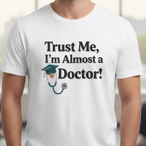 May include: White t-shirt with the text "Trust Me, I'm Almost a Doctor!" in black. A teal graduation cap and stethoscope graphic are below the text. The shirt is a crew neck and appears to be made of cotton.
