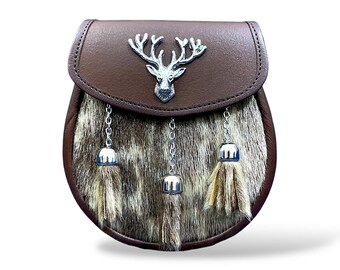 May include: A brown leather sporran with a silver stag head emblem on the flap. The sporran has a fur body and three silver tassel pendants with tan and brown fur. The sporran is a traditional Scottish accessory.