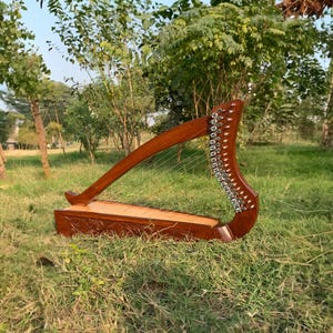 May include: A wooden harp with a rich, brown finish, resting on a bed of green grass. The instrument features a curved neck and a rectangular soundbox with carved details. The strings are visible, stretched across the frame.
