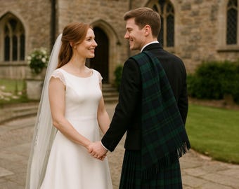 Puede incluir: Retrato de boda de una pareja tomados de la mano y sonriéndose. La novia lleva un vestido de novia blanco y un velo. El novio lleva un traje negro, un kilt escocés y una bufanda a juego. El fondo es un edificio de piedra.