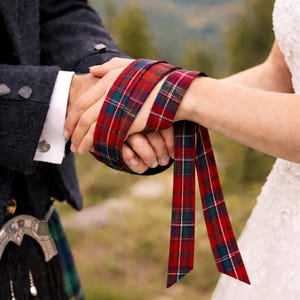 May include: A close-up of hands holding a red, green, and white plaid ribbon. The ribbon is wrapped around the wrists. The image suggests a wedding or commitment ceremony, with a blurred background of greenery.