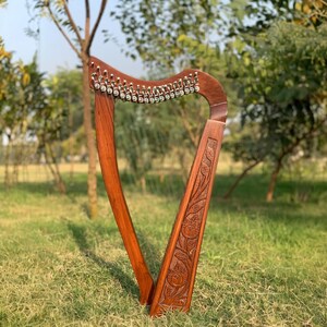 May include: A wooden harp with a rich, brown finish stands on a grassy surface. The instrument features a curved soundbox and a series of strings. Intricate floral carvings adorn the front of the harp, adding a decorative element to the musical instrument.