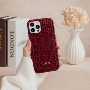 Puede incluir: Una funda de teléfono burdeos con textura de piel de cocodrilo y un bolsillo para tarjetas. La funda tiene las iniciales "JMN" en letras doradas. La funda del teléfono se sostiene en una mano.