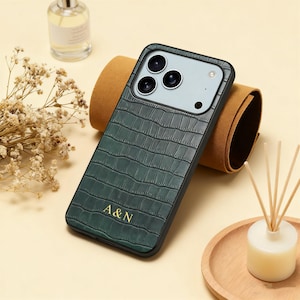May include: Dark green phone case with a crocodile skin texture and the gold letters "A&N". The case has a black border and a camera cutout. The phone case is resting on a brown surface.