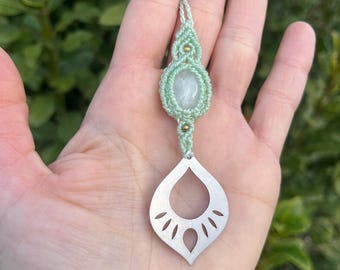 May include: A handmade macrame pendant necklace. The necklace features a light green cord, a central oval-shaped stone, and a silver-colored teardrop-shaped pendant with cut-out details. The pendant is held in a hand.