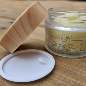 May include: A jar of tallow with a white lid and a bar of soap. The jar has a label with a cow illustration and the text "Stonetown Tallow". The soap is light brown with darker brown streaks. The items are on a wooden surface.