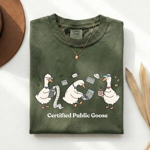 Könnte beinhalten: Olivgrünes T-Shirt mit einer Grafik von drei Comic-Gänsen. Eine Gans benutzt einen Taschenrechner, eine andere einen Laptop und die dritte hält eine Kaffeetasse. Der Text "Certified Public Goose" steht darunter.