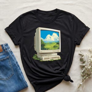 May include: Black t-shirt featuring a retro computer monitor graphic. The monitor displays a landscape scene with a blue sky, white clouds, and green fields. The shirt is paired with blue jeans and a small vase of white flowers.