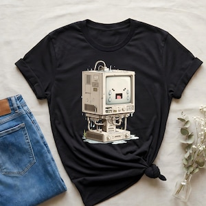 May include: Black t-shirt with a cartoon image of a retro computer with a crying face. The computer has a square screen and is on a base. The shirt is shown with blue jeans.