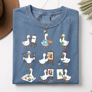 Puede incluir: Una camiseta azul con un diseño caprichoso de gansos de dibujos animados pintando. Los gansos se representan con pinceles, paletas y caballetes, creando obras de arte coloridas. La camiseta tiene un cuello redondo clásico y mangas cortas.