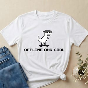 May include: White t-shirt featuring a pixelated dinosaur wearing sunglasses and riding a skateboard. The text "OFFLINE AND COOL" is printed below the graphic in a pixelated font. The shirt is paired with blue jeans.