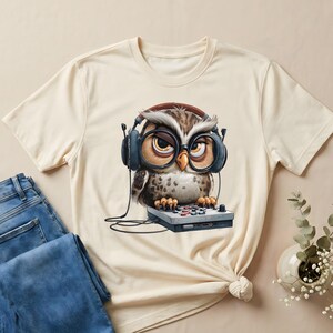 May include: Cream-colored t-shirt featuring a cartoon owl wearing headphones and glasses, perched on a small console. The owl has brown and white feathers and a focused expression. The shirt is paired with blue jeans.