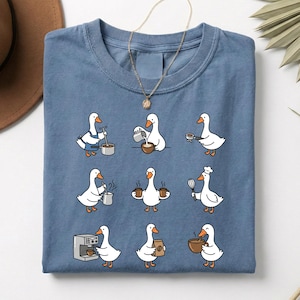 May include: A blue t-shirt featuring a repeating pattern of cartoon geese in various kitchen-related activities. The geese are depicted preparing coffee, wearing chef hats, and holding mugs. A gold necklace is draped over the shirt.