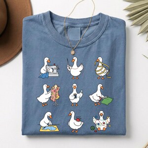 Puede incluir: Una camiseta azul con un diseño caprichoso de gansos involucrados en diversas actividades de costura. Los gansos se representan con máquinas de coser, cintas métricas y otros suministros de artesanía. Un collar dorado está drapeado sobre la camisa.