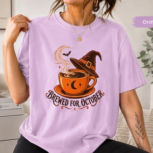 Op de afbeelding: Een lichtpaars t-shirt met een afbeelding van een oranje koffiekopje met een halve maan, een heksenhoed en opstijgende stoom. De tekst "BREWED FOR OCTOBER" staat in een donkerbruin lettertype onder het kopje, vergezeld door een kleine vleermuis.
