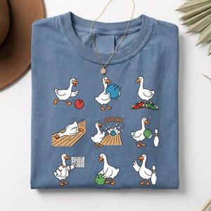 May include: A blue t-shirt featuring a whimsical design of cartoon geese playing bowling. The geese are depicted in various bowling scenarios, with bowling balls, pins, and a scoreboard. The word "STRIKE" is also visible on the shirt.