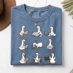 May include: A blue t-shirt featuring a whimsical design of cartoon geese engaged in various activities, such as using laptops, reading books, and dealing with tech issues. The geese are depicted in different poses, adding a playful touch to the shirt's design.