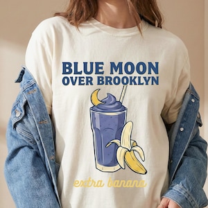 Puede incluir: Camiseta color crema con un gráfico de un batido azul, una luna creciente y un plátano. El texto "BLUE MOON OVER BROOKLYN" está encima del gráfico, y "extra banana" debajo.
