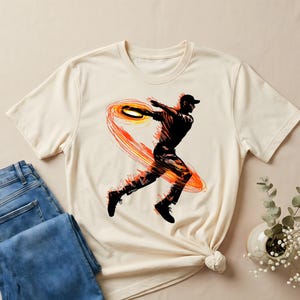 May include: A cream-coloured t-shirt with a graphic of a person throwing a disc, encircled by an orange and red design. The person is black, wearing a cap and trainers. Folded blue jeans are placed beside the shirt.