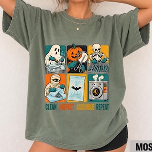 May include: A moss green short-sleeve t-shirt featuring a graphic print of six squares depicting a ghost, pumpkin, skeleton, mummy, bat, and washing machine. Below the graphics, the text reads CLEAN INSPECT ASSEMBLE REPEAT in bold letters.