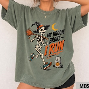 May include: A moss green t-shirt featuring a graphic of a skeleton wearing a witch hat and running with a broom. The text reads "MY BROOM BROKE I RUN" above a tombstone marked "1 MILE" with a small jack-o'-lantern illustration and a yellow crescent moon.