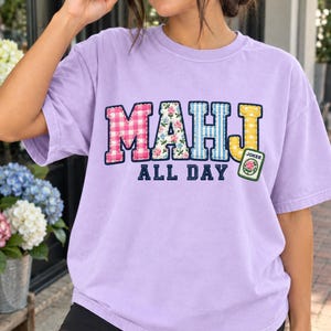 May include: A light purple short-sleeve t-shirt featuring the text MAHJ in large, multi-patterned letters with a pink gingham M, floral A, blue striped H, and yellow polka dot J. Below, the text ALL DAY appears in navy blue, with a Joker tile graphic.