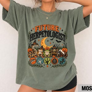 Puede incluir: Una camiseta verde musgo con el texto "FUTURE HERPETOLOGIST" en naranja y negro. El gráfico incluye ilustraciones de una serpiente, un gecko, una rana, una tortuga y un lagarto con sombreros de bruja, rodeados de calabazas, murciélagos y una luna.