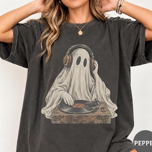 Könnte beinhalten: Ein anthrazitfarbenes Kurzarm-T-Shirt mit einem Grafikdruck eines weißen Geistes, der braune Kopfhörer trägt und einen braunen Plattenspieler bedient. Der Geist hat zwei ovale Augen und ist mittig auf der Vorderseite des Kleidungsstücks platziert.