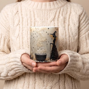 May include: A cylindrical candle featuring a black silhouette of a witch wearing a pointed hat and holding a staff. A black cauldron sits at the base with white smoke rising from it. The background is cream-colored with small black stars.