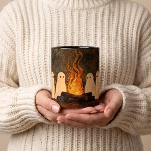 May include: A ceramic mug featuring a dark, forest-themed illustration of several white ghost figures sitting around a bright orange campfire with glowing flames and dark logs. The mug has a black interior rim and a smooth, cylindrical exterior surface.