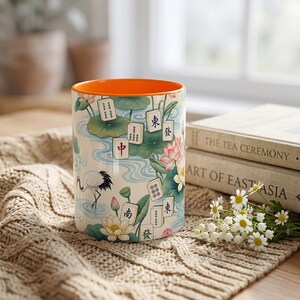 May include: A ceramic mug with a bright orange interior and a cream-colored exterior featuring a pattern of mahjong tiles, pink lotus flowers, green lily pads, and a white crane. The design includes blue water ripples and various Chinese characters.