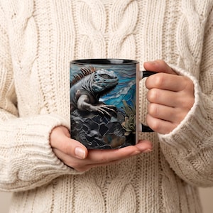 Op de afbeelding: Een keramische mok met een gedetailleerde grafische illustratie van een grijze leguaan op donkere rotsen tegen een blauw-witte achtergrond in glas-in-loodstijl. De mok heeft een zwart handvat en een zwarte binnenrand met een reptielontwerp.